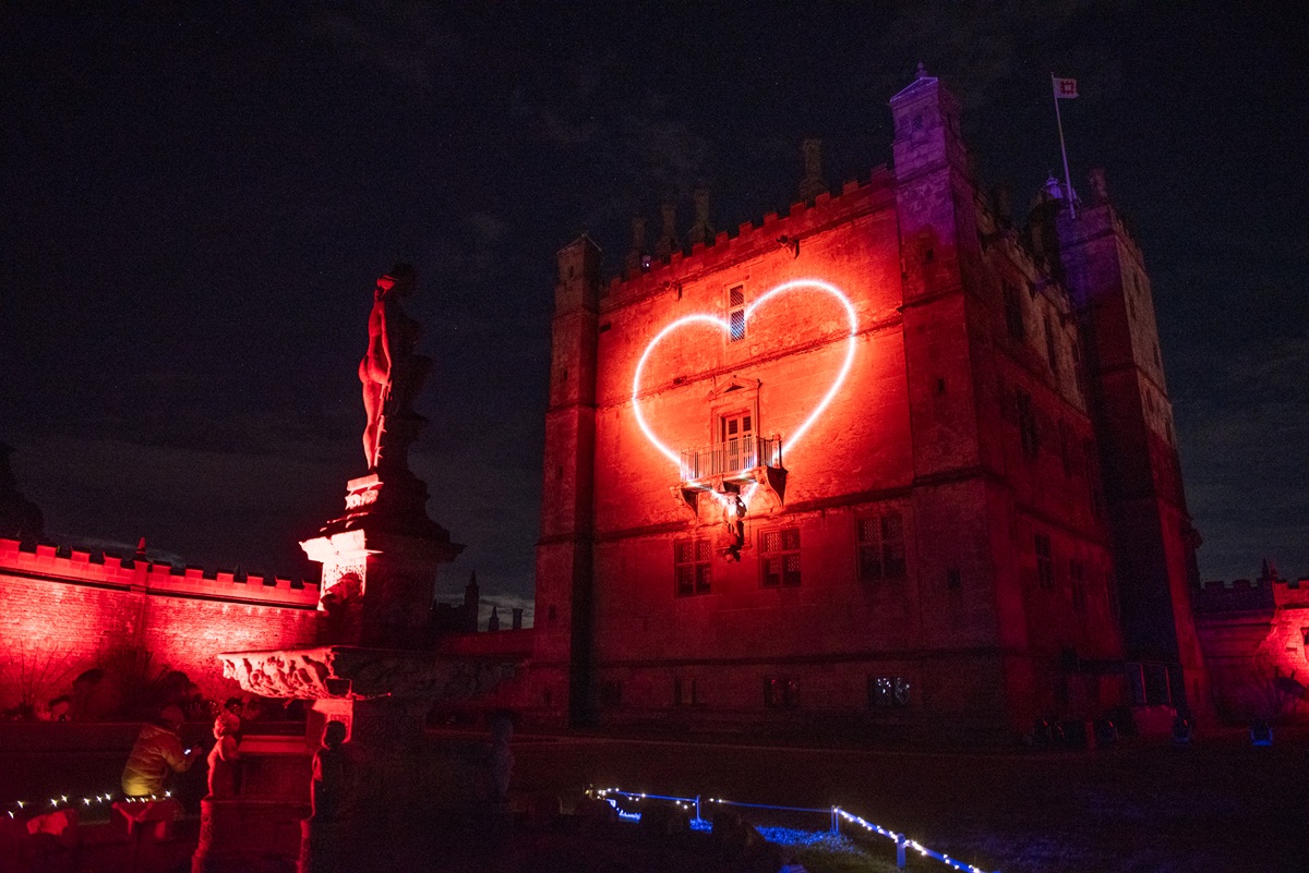 Large stone castle illuminated deep red at night, with a glowing neon-style heart projected onto its central wall. A statue on a plinth is lit red in the foreground, and a path edged with small white and blue lights leads toward the building under a dark, starry sky.